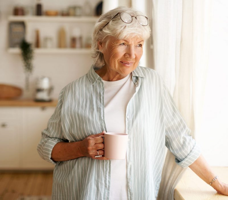 Coziness, domesticity and leisure concept. Portrait of stylish gray haired woman with round spectacles on her head enjoying morning coffee, holding mug, looking outside through window glass